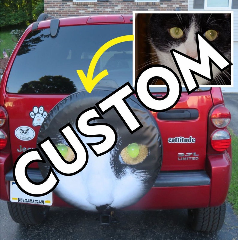May include: A red Jeep Liberty with a custom cat-themed spare tyre cover. The cover features a black and white cat with green eyes. The car has a "cattitude" sticker on the back.