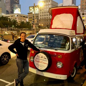 May include: A red and white vintage van with a sign that says "San Francisco's Hometown Creamery" and "Homemade Ice Cream & Desserts". The van is parked on a city street with a man standing next to it.