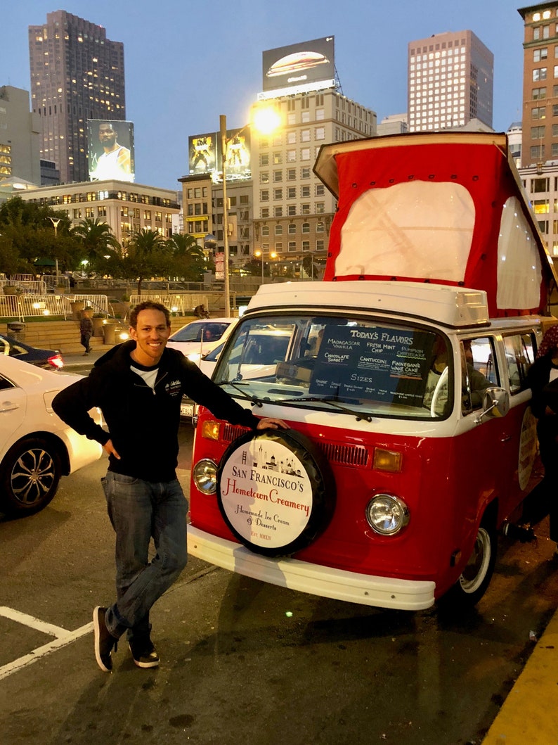 May include: A red and white vintage van with a sign that says "San Francisco's Hometown Creamery" and "Homemade Ice Cream & Desserts". The van is parked on a city street with a man standing next to it.