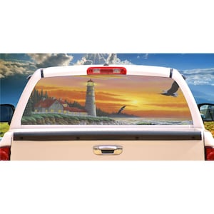 May include: A truck's rear window with a sunset scene decal. The decal features a lighthouse, a house, a bird flying, and the ocean. The sky is orange and yellow, and the water is blue.