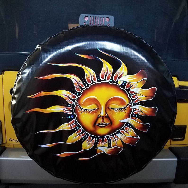 May include: A black spare tire cover with a yellow sun with a face and closed eyes. The sun has a yellow, orange, and red design. The background is black.