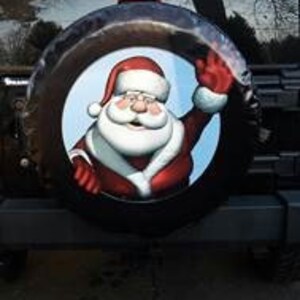 May include: A black spare tire cover with a festive Christmas design featuring a smiling Santa Claus waving. The background is blue with white clouds.
