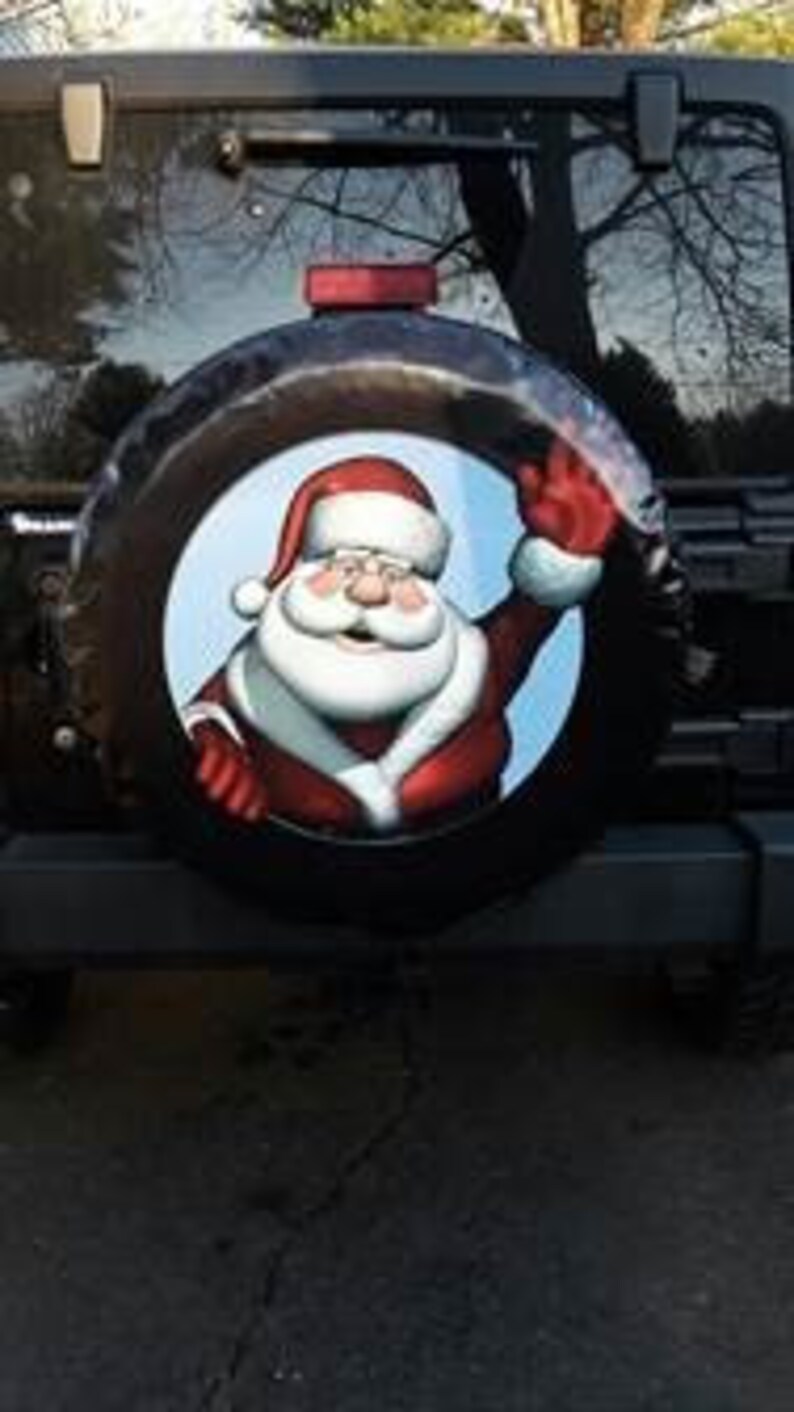 May include: A black spare tire cover with a festive Christmas design featuring a smiling Santa Claus waving. The background is blue with white clouds.