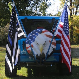 May include: A blue Jeep with a spare tire cover featuring an American flag design with an eagle. The Jeep has two flag poles with a black and white police flag and an American flag.