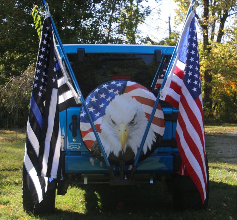 May include: A blue Jeep with a spare tire cover featuring an American flag design with an eagle. The Jeep has two flag poles with a black and white police flag and an American flag.