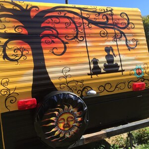 May include: A yellow camper van with a black and white painted design of a tree, a swing, and two figures sitting on the swing. The design also includes a peace sign and a sun with a face on the spare tire cover.