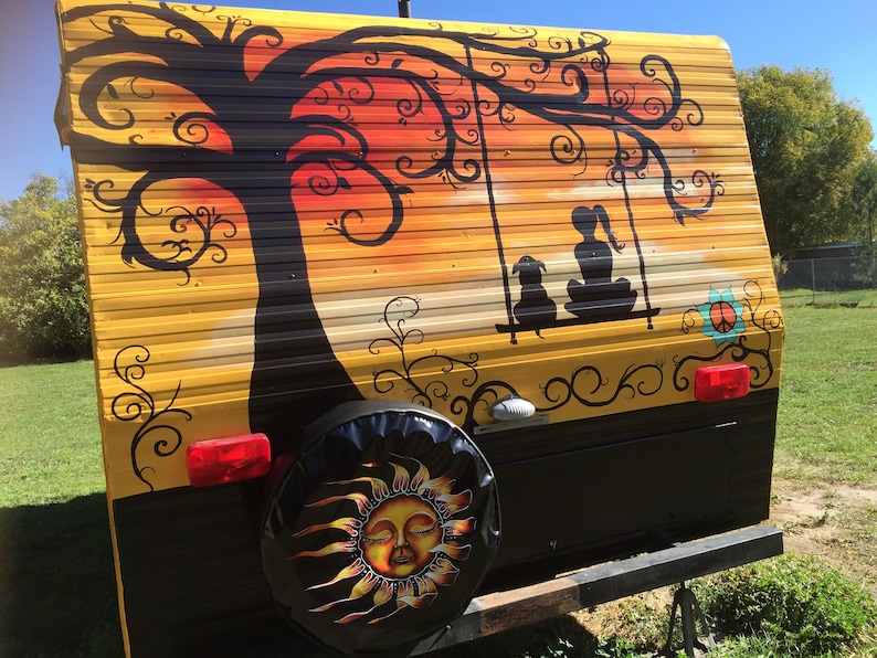 May include: A yellow camper van with a black and white painted design of a tree, a swing, and two figures sitting on the swing. The design also includes a peace sign and a sun with a face on the spare tire cover.
