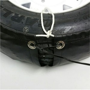 May include: A black tire cover with a white rope tied around it. The cover has two metal grommets with black straps attached to them. The straps are tied together with a black plastic tie.