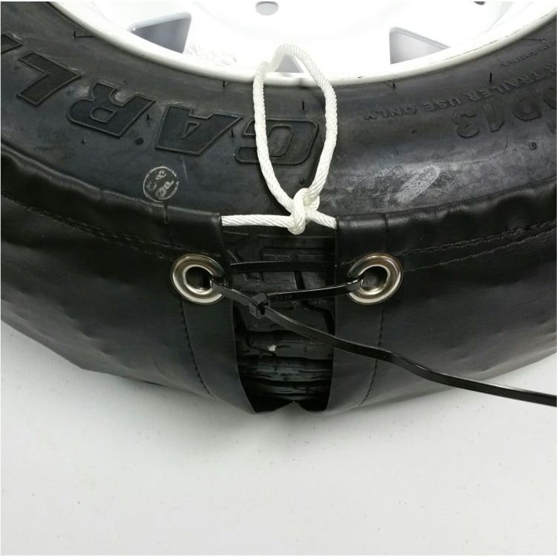 May include: A black tire cover with a white rope tied around it. The cover has two metal grommets with black straps attached to them. The straps are tied together with a black plastic tie.