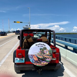 May include: A red Jeep driving on a bridge over a body of water. The spare tyre has a custom cover with the words "Matlacha est. 1926" and a colourful illustration of boats and buildings.
