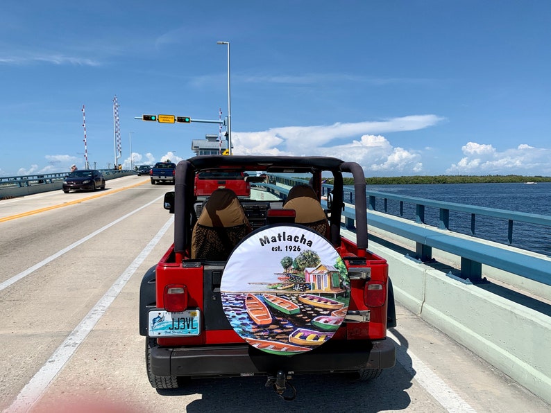 May include: A red Jeep driving on a bridge over a body of water. The spare tyre has a custom cover with the words "Matlacha est. 1926" and a colourful illustration of boats and buildings.