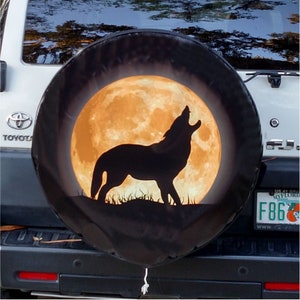 May include: A black spare tire cover with a howling wolf silhouette in front of a large, orange full moon. The cover is mounted on a white Toyota SUV.