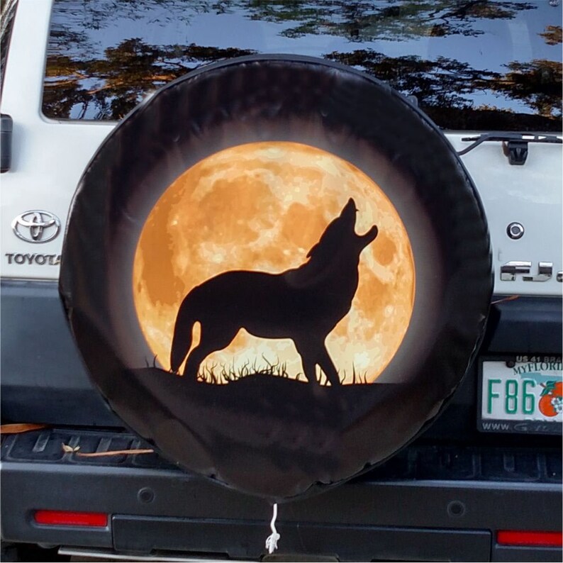 May include: A black spare tire cover with a howling wolf silhouette in front of a large, orange full moon. The cover is mounted on a white Toyota SUV.