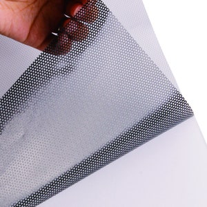 May include: A sheet of black and white perforated vinyl with a grid pattern. The vinyl is being peeled back to reveal the adhesive backing.