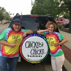 May include: Two people wearing tie-dye shirts stand in front of a black car. The car has a white round sign on the back that says "Chickpea Chicks Hummus ChickpeaChicks.com".