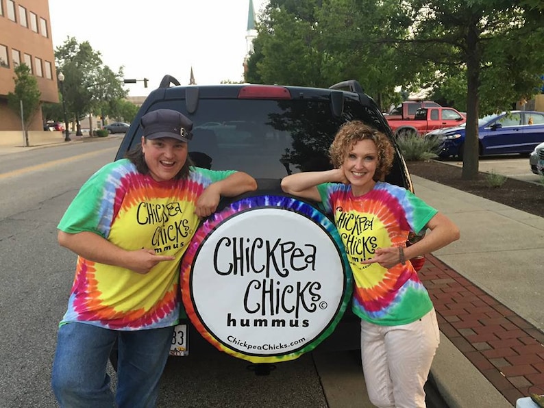 May include: Two people wearing tie-dye shirts stand in front of a black car. The car has a white round sign on the back that says "Chickpea Chicks Hummus ChickpeaChicks.com".