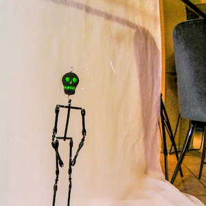 May include: A black skeleton puppet with a green skull head hanging from a black metal rod. The skeleton is made of wire and is posed in a standing position with its arms at its sides.