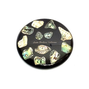 May include: A round black coaster with a design of iridescent abalone shell pieces. The coaster has the text "Santa Barbara, California" in white lettering.