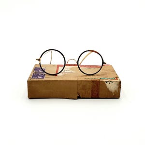 May include: Round, black-rimmed eyeglasses with gold-toned arms rest on a brown cardboard box. The box has red and white tape and blue stamps. The glasses have clear lenses.