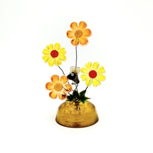 May include: A decorative glass flower arrangement featuring yellow and orange flowers with red centers, a small bee, and a gold-colored vase. The flowers are on thin black stems. The arrangement is set against a white background.