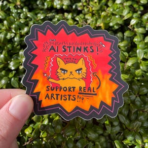 Könnte beinhalten: Ein roter und orangefarbener Aufkleber mit einer Comic-Katze, die einen Pinsel hält. Die Katze hat einen wütenden Ausdruck und der Text "AI STINKS!" und "SUPPORT REAL ARTISTS!" steht auf dem Aufkleber.