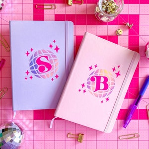 May include: Two pink and purple journals with a disco ball design and the letters "S" and "B" on the front. The journals are on a pink cutting mat with gold clips and silver disco balls.