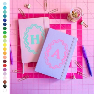 May include: Two pink and purple notebooks with a decorative border and the letters "H" and "A" in the center. The notebooks are on a pink cutting mat with gold clips and a jar of gold glitter.