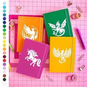 May include: Four colorful notebooks: orange, green, yellow, and magenta. Each notebook features a white mythical creature design: a phoenix, dragon, unicorn, and another phoenix. The notebooks are on a pink grid surface with office supplies.