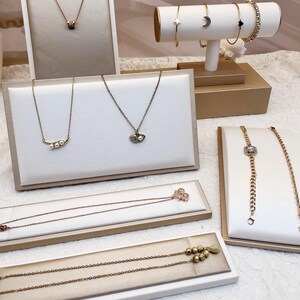 May include: A display of gold-toned jewelry, including necklaces and bracelets, presented on white and gold display stands. The necklaces feature various pendant designs, and the bracelets include star, moon, and heart motifs.