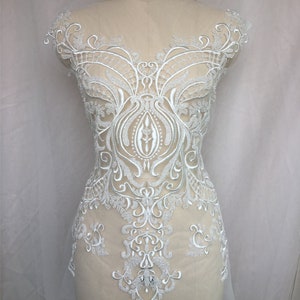 Może przedstawiać: Biała aplikacja z koronki o delikatnym wzorze kwiatowym. Koronka jest misterna i zawiera różne wzory, w tym spirale, liście i kwiaty. Aplikacja idealnie nadaje się do dodania odrobiny elegancji do każdego ubrania.