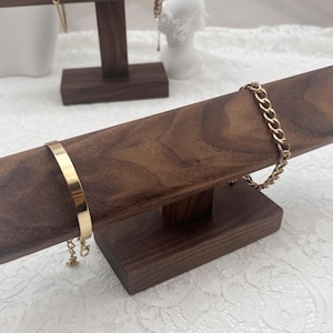 May include: A dark brown wooden T-bar jewelry display stand featuring a horizontal bar supported by a rectangular base. A smooth gold-tone cuff bracelet and a gold-tone chain link bracelet are draped over the horizontal wooden bar.
