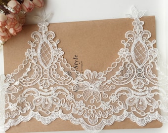 May include: White lace trim with floral and geometric patterns. The delicate lace features a scalloped edge and is displayed on a brown surface. The lace is likely used for embellishing garments or crafts.