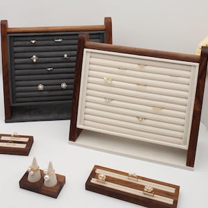May include: Two wooden ring display stands with dark brown frames and gray and cream-colored ring holders. Several rings with pearls and gemstones are displayed on the stands. Additional wooden ring holders are in the foreground.