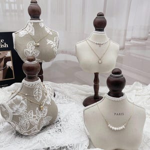 May include: Four jewelry display busts with wooden bases and tops. Two busts have a floral pattern, and two are plain. The busts display necklaces with pearls and pendants. One bust has the word "PARIS" on it.