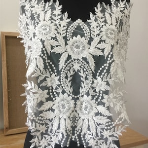 以下が含まれることがあります： パールビーズ付きの白い花柄レースアップリケ。レースは繊細な花柄のデザインで、複雑なディテールが施されています。このアップリケは、どんな衣服にもエレガントなタッチを加えるのに最適です。