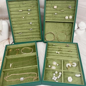 Könnte beinhalten: Vier grüne Schmuck-Präsentationsschalen mit Ringen, Armbändern, Halsketten und Ohrringen. Die Schalen haben ein weiches, grünes Samtfutter. Der Schmuck umfasst Gold- und Silberstücke mit Perlen- und Blumenakzenten. Die Schalen sind auf einer weißen Oberfläche angeordnet.