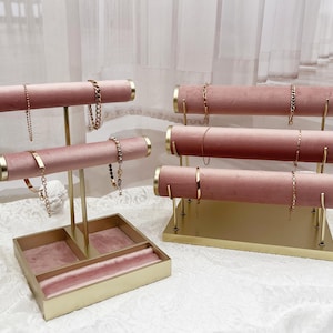 May include: Pink velvet jewellery display stands with gold accents. The stands are designed to hold bracelets and necklaces. One stand has a built-in tray for additional jewellery storage. The stands are on a white lace surface.