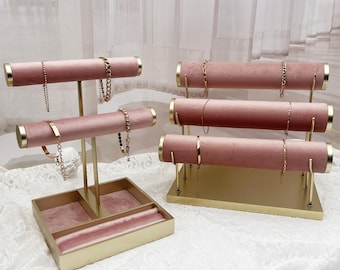 May include: Two jewelry display stands featuring gold-toned metal frames and soft pink velvet-covered cylindrical bars. The left stand has two tiers and a square base with compartments, while the right stand has three tiers on a rectangular base.