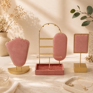 Op de afbeelding: Een verzameling goudkleurige sieraden displays met roze fluwelen inzetstukken. De displays omvatten een kettingstandaard, een oorringhouder en een kleine rechthoekige display. De sieradenhouders staan op een lichtgekleurd oppervlak.