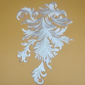 May include: White embroidered floral applique with a detailed, feathery design. The applique features a symmetrical, stylized leaf pattern, perfect for embellishing garments or crafts. The design is set against a yellow background.