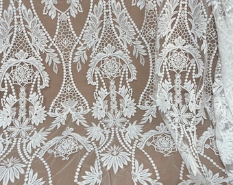 May include: White lace fabric with an intricate floral and leaf pattern. The design features detailed embroidery with a repeating motif of flowers, leaves, and geometric shapes. The fabric is semi-sheer, with a brown background visible through the openwork.