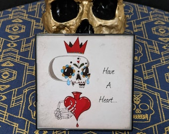 May include: A square framed print with a white background featuring a sugar skull wearing a red crown and holding a red heart with the text "Have A Heart..."