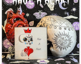 May include: A white ceramic skull with a black and white sun design, a red heart, and a sugar skull with a crown and the text "Have A Heart..." on a white background.