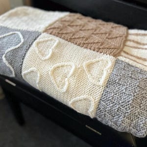 Könnte beinhalten: Eine handgestrickte Decke mit Patchwork-Design. Die Decke hat Quadrate in verschiedenen Texturen und Farben, darunter Creme, Grau und Braun. Ein Quadrat hat herzförmige Muster. Die Decke ist über einer schwarzen Oberfläche drapiert.
