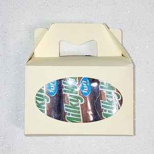 Puede incluir: Una caja de cartón blanca con asa y una ventana ovalada transparente. La ventana muestra un paquete de barras de chocolate Milky Way con el texto "fun size" en el envoltorio.