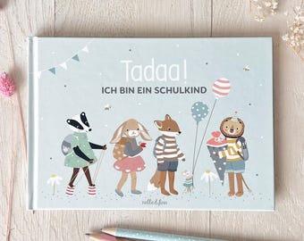 May include: A light blue hardcover book with the text "Tadaa! ICH BIN EIN SCHULKIND" and illustrations of animal characters. The cover features a badger, rabbit, fox, and lion with school supplies and balloons. Two pencils are in the foreground.