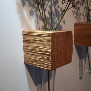 Könnte beinhalten: Zwei Wandvasen aus Holz mit klaren Glasröhren. Die Vasen sind aus Naturholz mit rustikalem Finish. In den Vasen befinden sich rosa und weiße Blumen.