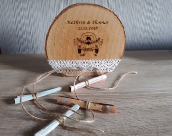 Puede incluir: Una rodaja de madera con un borde de encaje blanco y una inscripción personalizada que dice "Kathrin & Thomas 21.12.2018". Un pequeño coche de dibujos animados con una novia y un novio dentro está grabado en la madera. Cuatro pequeños trozos de papel enrollados están atados con cuerda y esparcidos sobre una superficie de madera.