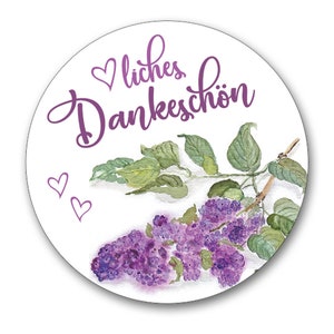May include: A round sticker with a purple watercolor illustration of lilac flowers and leaves. The text "liches Dankesch&ouml;n" is written in purple cursive lettering with a small heart above the word "liches".