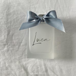 May include: A clear glass bottle with a silver lid and a light blue bow. The bottle has a white label with the name "Luca" written in black cursive.